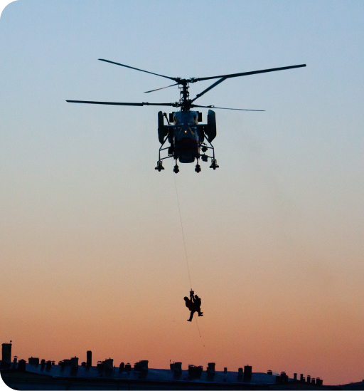 A helicopter in mid-air with a sunset in the distance showing 2 people hanging suspended on a line beneath it.